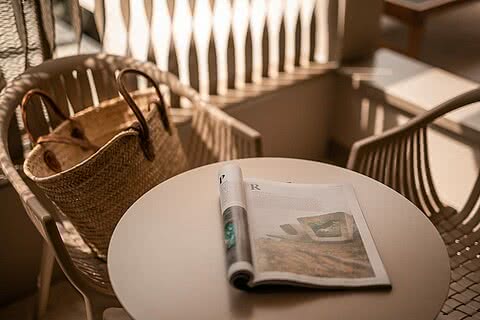 Balkon im Sonnenresort Wellnesshotel in Südtirol mit Tisch, Stühlen, einer geflochtenen Tasche und einer offenen Zeitschrift.